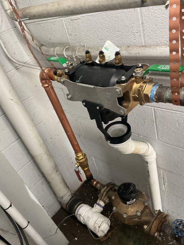 Installed reduced pressure zone (RPZ) backflow prevention assembly on commercial plumbing system per permit requirements requested 9/3. Assembly features brass fittings, test cocks, and green-handled isolation valves with copper and PEX piping connections mounted to wall in mechanical room, with white PVC drainage piping configured below the device.