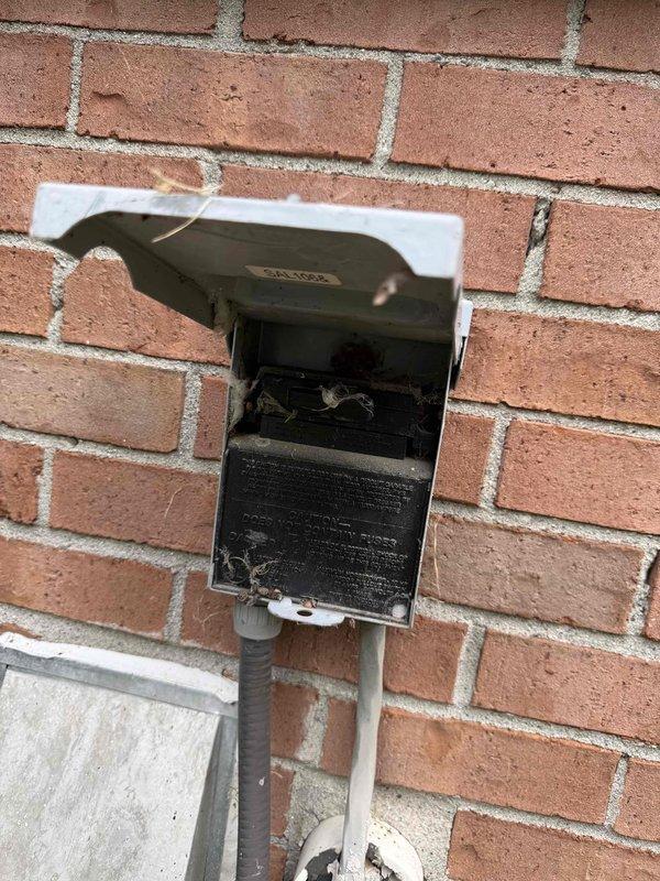 # HVAC Service Completed

Responded to no-cooling complaint affecting entire residence with 25-year-old air conditioning system. Condenser unit located at rear exterior of property.

Upon arrival, performed visual inspection of outdoor condensing unit and associated electrical components. Discovered severe fire damage to exterior electrical disconnect box mounted on brick wall near condenser. Interior of disconnect showed extensive charring, burned components, and heat damage to internal surfaces. Damage indicated electrical failure or short circuit event that compromised power supply to AC system.

Used digital multimeter to verify electrical readings and confirm loss of power at disconnect location. Tested voltage at various points in electrical circuit to isolate failure point. Black test leads documented voltage measurements at damaged disconnect box.

Photographed damaged electrical disconnect box showing burned interior components and compromised enclosure. Documented condition o