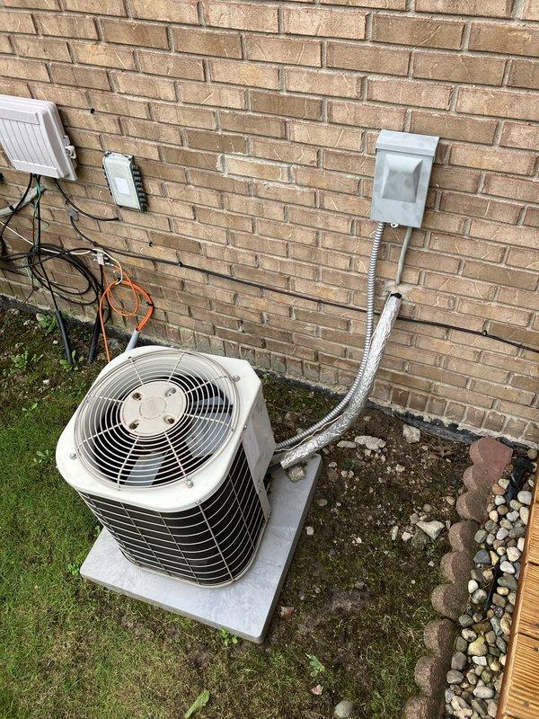 Completed full HVAC system estimate for residential property requiring both air conditioning and furnace replacement. Evaluated existing outdoor installation site against tan brick wall with adequate clearance and proper concrete pad foundation. Assessed electrical infrastructure including existing disconnect box, junction boxes, and conduit routing along exterior wall. Documented current equipment positioning, refrigerant line routing with insulation, and electrical service connections to ensure accurate sizing and installation planning for new system. Photographed installation area including ground conditions with grass and decorative rock border, deck proximity, and mounting surface to facilitate proper equipment selection and installation specifications for complete heating and cooling system replacement.
