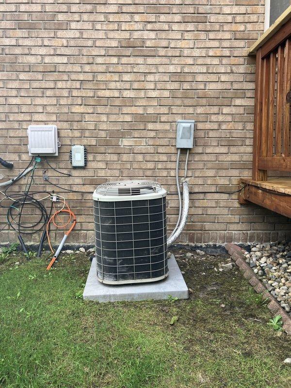 Completed full HVAC system estimate for residential property requiring both air conditioning and furnace replacement. Evaluated existing outdoor installation site against tan brick wall with adequate clearance and proper concrete pad foundation. Assessed electrical infrastructure including existing disconnect box, junction boxes, and conduit routing along exterior wall. Documented current equipment positioning, refrigerant line routing with insulation, and electrical service connections to ensure accurate sizing and installation planning for new system. Photographed installation area including ground conditions with grass and decorative rock border, deck proximity, and mounting surface to facilitate proper equipment selection and installation specifications for complete heating and cooling system replacement.