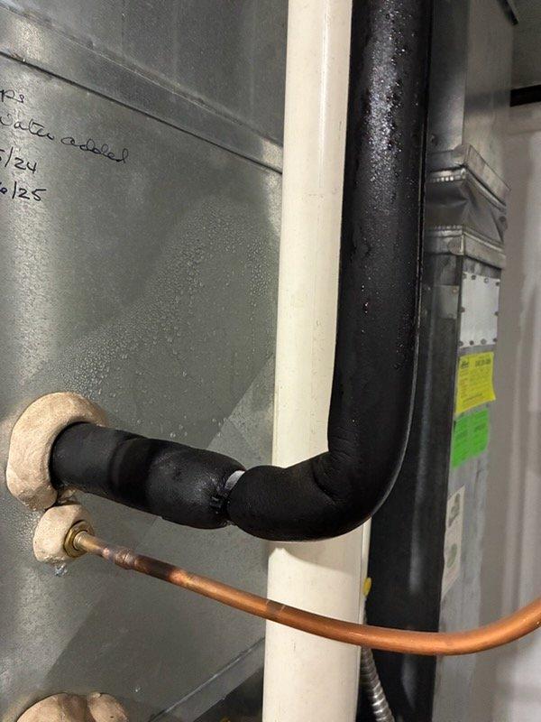 Responded to service call for reported condensation pump leak on basement cooling system. Upon arrival after 3PM as requested, inspected HVAC equipment and identified significant condensation accumulation on insulated refrigerant line and associated plumbing connections. Documented extensive moisture beading on black insulated suction line elbow joint, indicating proper system operation but potential drainage issue. Examined condensate removal system including PVC drain lines and pump assembly. Observed water damage and staining on floor surface beneath equipment, confirming active leak condition. Photographed affected areas showing copper refrigerant lines, brass fittings, and air handler connections for documentation purposes. Assessed condensate pump operation and drainage pathway to determine source of leak and recommend appropriate repairs.