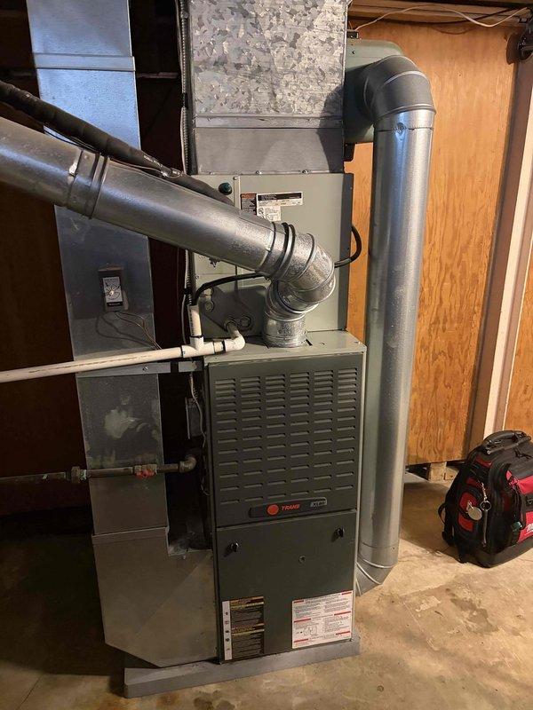 Responded to service call for reported leak originating from furnace system. Upon inspection of the basement-installed Trane forced air furnace (10+ years old), evaluated all visible plumbing connections, condensate drain lines, and PVC discharge piping for signs of active leakage or moisture accumulation. Examined the white PVC condensate drain line extending from the furnace, inspecting all joints, connections, and the floor-mounted base assembly for proper seal integrity. Assessed the condition of all ductwork connections, including the vertical cylindrical supply duct and return air plenum attachments, to rule out secondary moisture sources. Verified proper drainage function and confirmed no obstructions in the condensate removal system. Documented equipment condition and piping configuration with photographic evidence for service records.