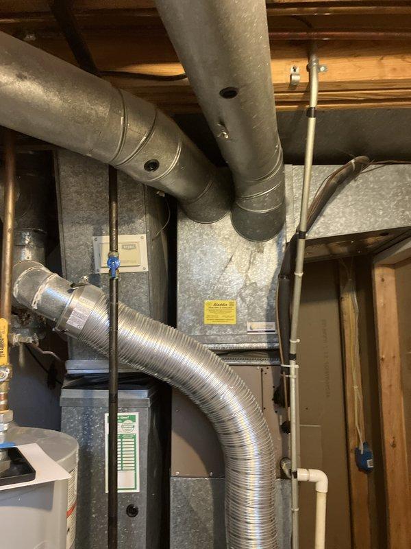 Conducted comprehensive assessment of existing basement HVAC system for replacement estimate on small residential property under 1500 square feet. Evaluated current electric heat pump system including air handler unit mounted in basement mechanical room with existing ductwork configuration. Documented equipment specifications, installation conditions, and spatial requirements for replacement system. Assessed metallic supply ductwork runs, flexible duct connections, and overall distribution system integrity. Photographed existing HVAC equipment showing gray metal air handler with visible manufacturer labels and warning placards, along with adjacent water heater and plumbing infrastructure. Evaluated basement installation environment including exposed ceiling joists, reflective insulation on walls, structural framing, and available clearances for new equipment installation. Prepared technical documentation and recommendations for complete heating and cooling system replacement compatible