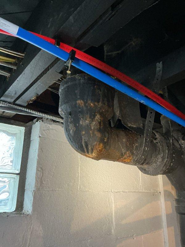 Responded to a water leak originating from the main drain pipe located beneath the toilet. Upon inspection, discovered severely corroded cast iron drain piping in the basement ceiling area showing extensive rust deterioration and active moisture damage. Removed floor tiles in the bathroom to access the toilet flange and underlying drain connection, exposing the compromised pipe section. Documented the extent of corrosion on the main drain stack and assessed the structural integrity of surrounding plumbing components, including the hot and cold water supply lines running adjacent to the affected area.