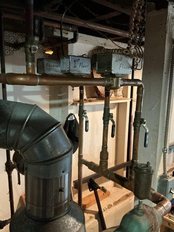 Provided estimate for hot water boiler replacement in basement mechanical room. Existing system is very old with malfunctioning controls that prevent proper shutdown, presenting safety and efficiency concerns. Current installation includes aged copper piping with significant patina, multi-zone heating distribution system with floor-designated controls, expansion tank, and circulation pumps showing considerable wear from years of operation.