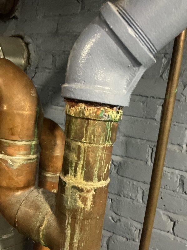 Completed comprehensive inspection on 18+ year old boiler system located in basement and evaluated secondary unit on back porch. Boiler system showed significant signs of age with extensive corrosion and verdigris buildup on copper piping throughout the mechanical room, indicating prolonged moisture exposure and material degradation. Documented deteriorated copper pipes with yellow-green patina, rust deposits, and oxidation at multiple connection points. Observed previous repair work where gray PVC components had been integrated with existing copper piping system. Inspected gate valves, pipe fittings, and ductwork connections on main heating unit. Back porch unit assessed separately and confirmed to be independent from main boiler system. Recommended monitoring corroded piping for potential leaks and future replacement consideration due to advanced deterioration of copper components.