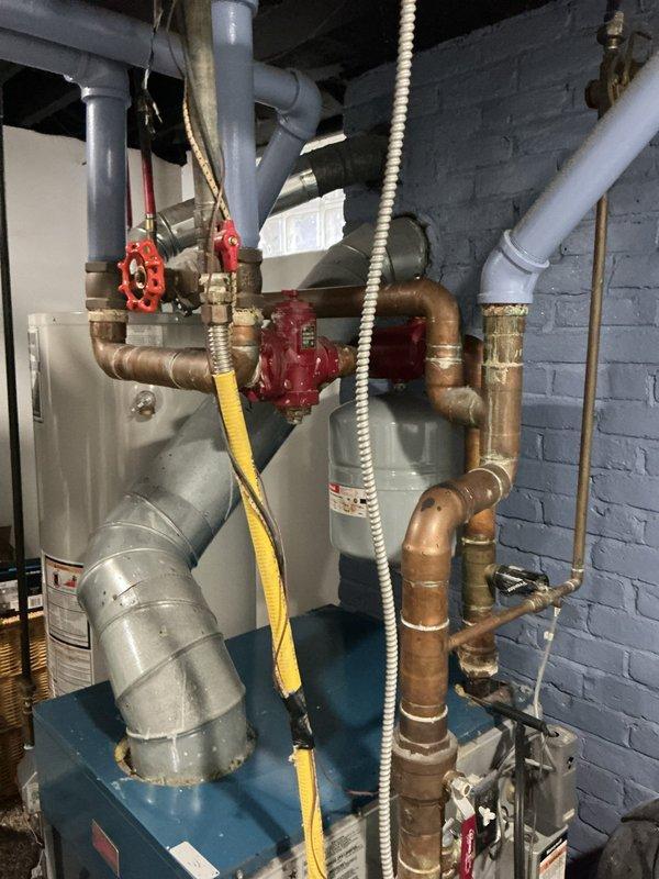 Completed comprehensive inspection on 18+ year old boiler system located in basement and evaluated secondary unit on back porch. Boiler system showed significant signs of age with extensive corrosion and verdigris buildup on copper piping throughout the mechanical room, indicating prolonged moisture exposure and material degradation. Documented deteriorated copper pipes with yellow-green patina, rust deposits, and oxidation at multiple connection points. Observed previous repair work where gray PVC components had been integrated with existing copper piping system. Inspected gate valves, pipe fittings, and ductwork connections on main heating unit. Back porch unit assessed separately and confirmed to be independent from main boiler system. Recommended monitoring corroded piping for potential leaks and future replacement consideration due to advanced deterioration of copper components.