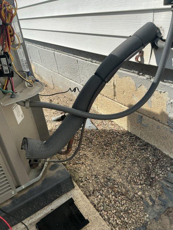 Responded to service call for 2-year-old forced air AC system in basement producing loud humming noise and green fluid leak from piping. Created 90-degree bends in refrigerant line set to proper routing configuration. Inspected indoor evaporator coil assembly and PVC condensate drain system where green-tinted condensate was discharging through drain line with red cap fitting. Examined outdoor condensing unit on concrete pad with insulated refrigerant lines entering building through exterior wall. Verified proper line set insulation installation on copper refrigerant piping and confirmed system connections at both indoor and outdoor units. Addressed refrigerant line routing concerns and resolved drainage issue.