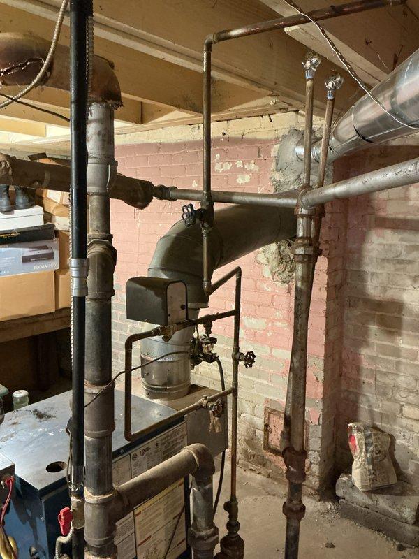 Conducted comprehensive boiler replacement estimate for aging 20-year-old heating system located in basement utility area. Evaluated existing dark green boiler unit with extensive piping infrastructure, including multiple steel supply and return lines, valves, and fittings showing age-related deterioration and surface corrosion. Assessed surrounding mechanical systems including overhead ductwork, exposed plumbing infrastructure, and electrical connections. Documented current equipment condition through photographic survey, noting worn components and installation configuration within basement space featuring pink-painted brick walls and concrete flooring. Provided detailed replacement recommendations and specifications for new boiler system suitable for property heating requirements.