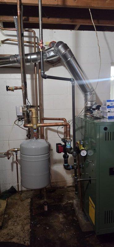 Completed full boiler installation in basement mechanical room, including green residential boiler unit with associated copper piping network, expansion tank, pressure gauges, brass valves, and metallic exhaust venting system. Installation included three thermostats and hydronic heating system connections to existing water heater. Mechanical, boiler, and reconnection permits were requested, with electrical contractor scheduled to return for final boiler wiring to complete the installation.