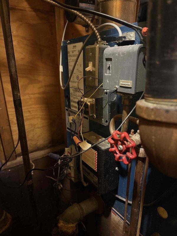 Responded to service call for steam boiler system in basement following report from plumbing contractor regarding multiple issues and leaks. Upon inspection, confirmed significant corrosion and deterioration on copper piping with heavy mineral deposits and rust buildup indicating active leak points. Examined blue vertical boiler unit with Honeywell control system and associated piping connections. Documented extensive pipe corrosion, water staining on surrounding wooden structural members, and compromised integrity of copper supply lines. Photographed corroded sections showing white mineral scaling and orange-brown oxidation on piping throughout the system. Advised homeowner of findings and recommended comprehensive evaluation for pipe replacement and potential boiler system repairs to address reported leaks and prevent further water damage to structure.