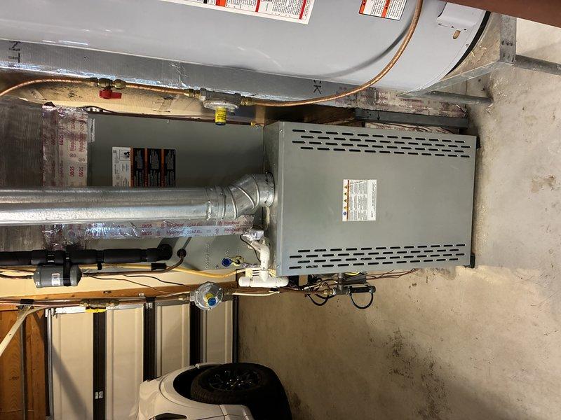 Completed Service Agreement Visit inspection of residential HVAC system. Documented basement furnace installation including gray metal furnace unit with proper ventilation, metallic ductwork with reflective insulation, and copper refrigerant line connections. Photographed outdoor condensing unit from above, noting black circular fan motor assembly and gray drain pan with standing water and visible algae growth indicating drainage concerns that may require attention during future maintenance visits.