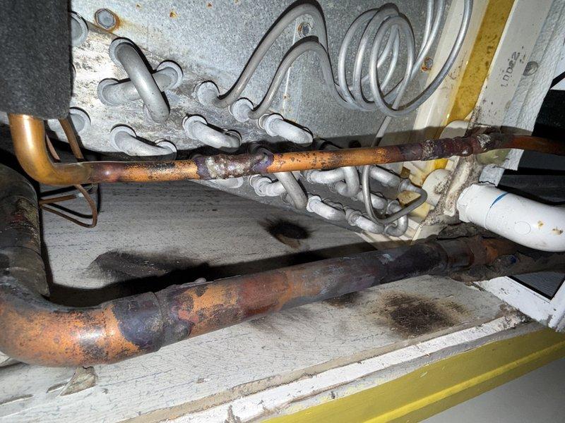 # Turn Over Equipment - Leaky Evaporator

Responded to service call for equipment turnover involving a leaking evaporator coil. Upon arrival, performed comprehensive inspection of the air handler unit and associated refrigeration components. Found evidence of moisture accumulation and condensation on the galvanized metal casing of the air handler, indicating active refrigerant leak from the evaporator coil.

Documented extensive corrosion and deterioration on multiple copper refrigerant lines throughout the system. The horizontal suction line exhibited severe oxidation with heavy rust buildup, compromising system integrity. Inspected electrical components including capacitor, contactors, relays, and associated wiring - all appeared functional but system could not operate safely with compromised evaporator coil.

Photographed the air handler unit showing moisture streaking on exterior surfaces, exposed blower assembly, and electrical connections. Captured detailed images of the severely