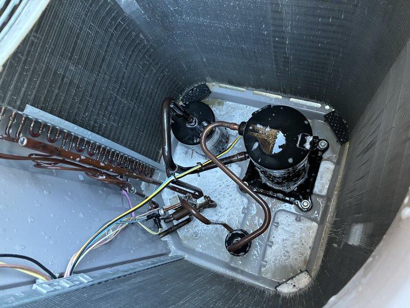Completed first service agreement visit on heating system with UV light under 12-month warranty. Upon inspection, discovered significant frost and ice buildup throughout the refrigeration compartment, coating the evaporator coil, compressor unit, and base surfaces. Photographed the cylindrical HVAC unit interior showing the copper evaporator coil with parallel fins, black hermetic compressor with copper refrigerant line connections, and electrical wiring. Documented the white condensate drain pan with center-mounted drain fitting assembly, copper piping with metal brackets, and moisture accumulation on pan surface. Verified proper installation of condensate pump with electrical connections. System components including evaporator coil, compressor, refrigerant lines, and drainage system were assessed for proper operation under warranty coverage.
