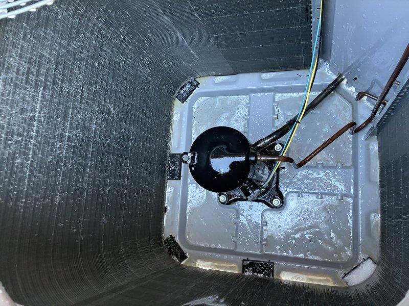 Completed first service agreement visit on heating system with UV light under 12-month warranty. Upon inspection, discovered significant frost and ice buildup throughout the refrigeration compartment, coating the evaporator coil, compressor unit, and base surfaces. Photographed the cylindrical HVAC unit interior showing the copper evaporator coil with parallel fins, black hermetic compressor with copper refrigerant line connections, and electrical wiring. Documented the white condensate drain pan with center-mounted drain fitting assembly, copper piping with metal brackets, and moisture accumulation on pan surface. Verified proper installation of condensate pump with electrical connections. System components including evaporator coil, compressor, refrigerant lines, and drainage system were assessed for proper operation under warranty coverage.