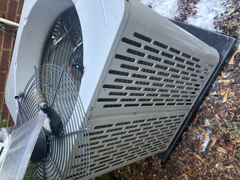 Performed a first service agreement visit on the heat pump system, including inspection of the outdoor condenser unit with the top section removed for access to the fan assembly, condenser coil, compressor, and refrigerant line set. Cleaned the condenser coil with a brush to remove debris and restore airflow, and noted frost accumulation around the unit consistent with cold-weather heat pump operation. Observed oily residue pooled on the compressor base plate near the refrigerant fittings, indicating a possible refrigerant or oil leak that warrants further leak testing and evaluation.
