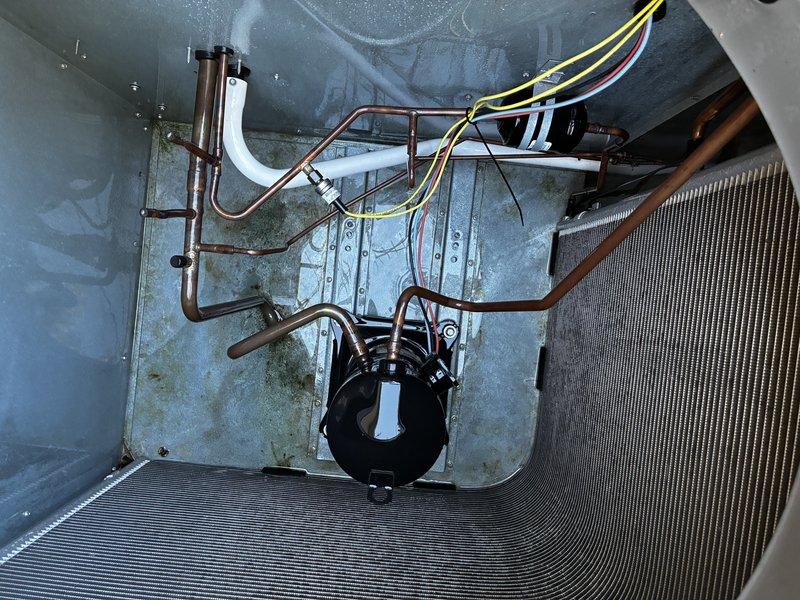 Performed a scheduled maintenance inspection on the packaged HVAC unit, including a visual check of the cabinet, electrical disconnect, conduit, and condensate drain line, all found intact with no signs of rust, frost, or damage. Opened the unit to inspect the compressor, refrigerant lines, capacitor, wiring, and condenser coil fins, noting algae and staining on the base pan and a loose zip tie near the wiring harness. Recommend maintaining clearance around the unit by trimming the surrounding shrubs and grasses to ensure proper airflow.