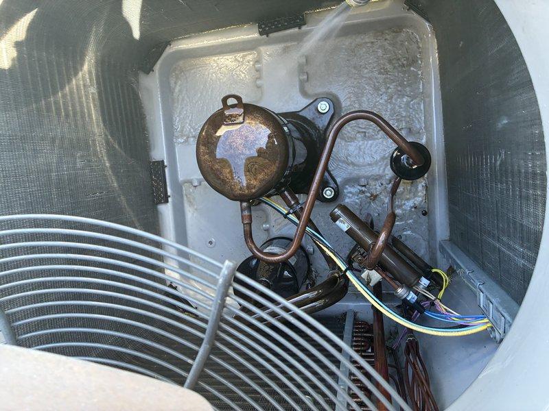 Performed a service agreement visit on the outdoor condenser unit, removing the fan assembly and guard to access the interior components. Inspected the condenser coil fins, compressor, refrigerant line set, capacitor, and electrical wiring and terminals, and washed the coil and cabinet to clear accumulated dirt and debris. Noted surface corrosion on the compressor shell and moisture and frost-like buildup on the base pan, which should be monitored at future visits.