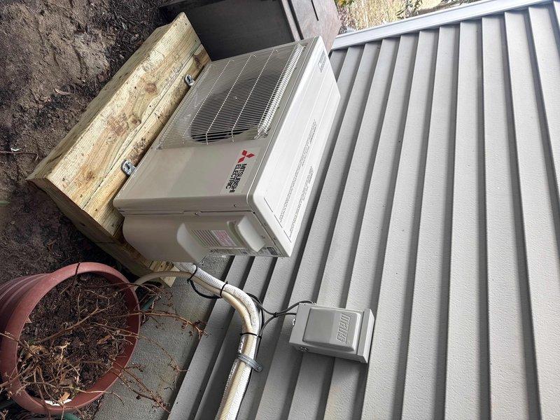 Removed old geothermal split system from bonus room kneewall location and installed new Mitsubishi ductless system consisting of MUZ-GX12NL 12,000 BTU 25 SEER outdoor heat pump unit and MSZ-GX12NL wall-mounted indoor air handler with remote thermostat. Positioned outdoor unit on custom wooden stand near crawlspace entrance, reused and extended existing disconnect, and ran new lineset following previous geothermal split system pathway.