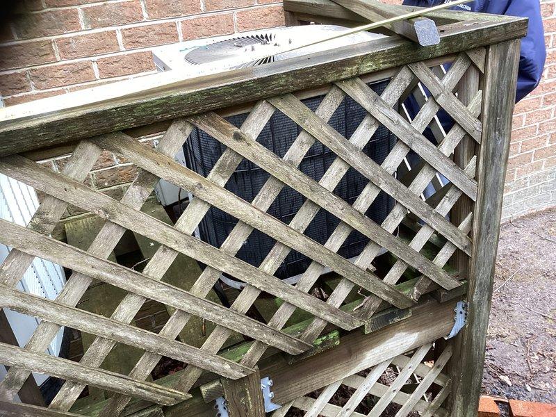 Conducted inspection of non-operational 23-year-old HVAC system located in attic. Assessed two condenser units against brick exterior wall, both showing significant age deterioration with weathered housings and accumulated debris. Wooden lattice enclosure surrounding units exhibits substantial weathering and mildew growth, requiring replacement along with the complete HVAC system.