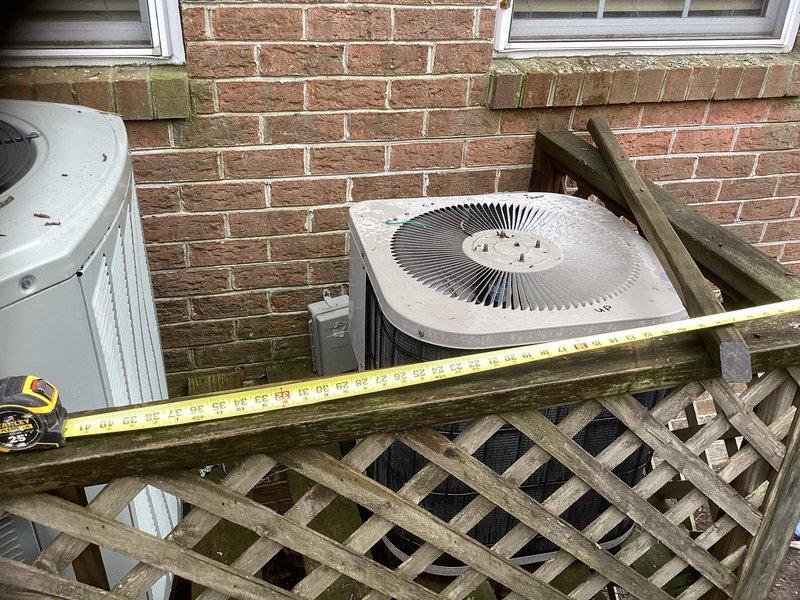 Conducted inspection of non-operational 23-year-old HVAC system located in attic. Assessed two condenser units against brick exterior wall, both showing significant age deterioration with weathered housings and accumulated debris. Wooden lattice enclosure surrounding units exhibits substantial weathering and mildew growth, requiring replacement along with the complete HVAC system.