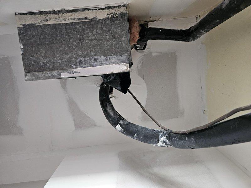 Relocated and reinstalled air handler unit in closet following completion of plumbing work. Cut out drywall ceiling section to accommodate repositioning of upstairs refrigerant lines, then covered opening with flat metal panel above duct fittings. Installed plywood base under existing return plenum to prepare for future flooring installation. Repositioned air handler to right side of closet with service panels facing left to create clearance in recessed area. Relocated lineset to back wall of closet to accommodate new air handler placement. Installed new electrical disconnect on left side of closet and ran power wire across interior closet wall above door to air handler connection point.