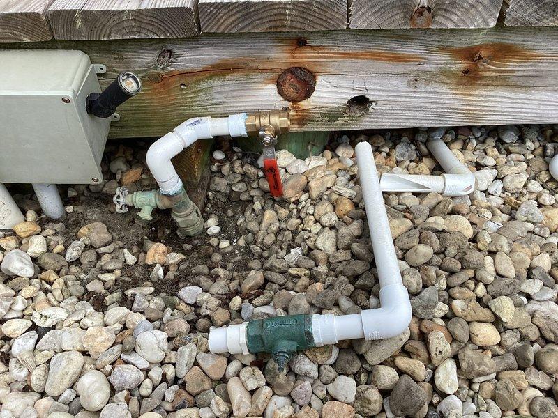 Completed de-winterization service for residential property with multiple outdoor water fixtures. Restored two outdoor shower systems to operational status by activating water supply lines, opening main shutoff valves, and pressurizing the plumbing system. Inspected rooftop plumbing assembly showing significant weathering and corrosion on copper components with verdigris oxidation present on base sections. Examined ground-level PVC distribution system beneath deck structure, verified proper operation of brass ball valves and control points, and confirmed water flow through all connection points. All outdoor water fixtures tested and confirmed operational for seasonal use.