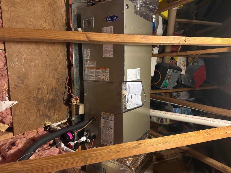 Completed full HVAC system replacement including removal and disposal of existing equipment. Installed new Carrier 27SCA524A003 2-ton 15 SEER heat pump on pressure-treated wood platform with Carrier FJ5ANXB24L00 air handler and 10kW electric heat pack in basement utility area. Fabricated and installed metal duct transitions to adapt new equipment to existing ductwork, reused existing condensate drain lines, replaced outdoor disconnect and electrical whip, and installed 14x24x1 filter grille and Honeywell TH6320WF digital WiFi thermostat for system control.