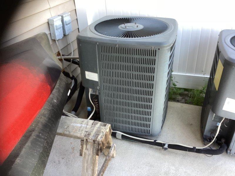 Assessed 19-year-old Goodman air conditioning system utilizing R-22 refrigerant and confirmed low refrigerant charge. Documented existing outdoor condenser unit installation with photographs showing multi-unit configuration on concrete pad with electrical disconnect and refrigerant line connections. Provided customer with replacement estimates for AC-only replacement option as well as complete HVAC system replacement option.