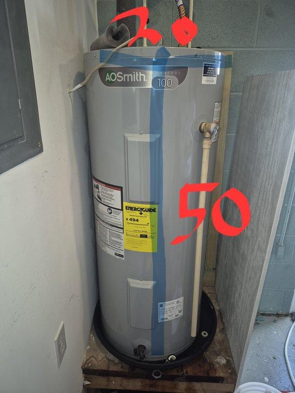 Responded to emergency call for leaking water heater in older residential property. Upon arrival, inspected AO Smith model 100 tanked water heater showing signs of active leak. Unit was installed in utility room on concrete floor with standard copper plumbing connections and proper venting. Assessed the condition of the approximately 50-gallon capacity water heater, noting the age and wear of the equipment consistent with the home's 1960s construction era. Documented the installation setup including mounting platform, piping connections, brass fittings, and overflow pan configuration. Photographed the unit from multiple angles to record the leak source, overall equipment condition, and surrounding installation environment for service records and customer reference.