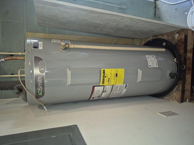 Responded to emergency call for leaking water heater in older residential property. Upon arrival, inspected AO Smith model 100 tanked water heater showing signs of active leak. Unit was installed in utility room on concrete floor with standard copper plumbing connections and proper venting. Assessed the condition of the approximately 50-gallon capacity water heater, noting the age and wear of the equipment consistent with the home's 1960s construction era. Documented the installation setup including mounting platform, piping connections, brass fittings, and overflow pan configuration. Photographed the unit from multiple angles to record the leak source, overall equipment condition, and surrounding installation environment for service records and customer reference.