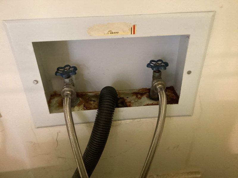 Responded to water leak complaint with visible ceiling damage in dining room below bathroom and laundry area. Relocated washing machine to access wall-mounted supply valves through access panel. Inspection revealed significant rust staining and corrosion around water supply connections, indicating chronic moisture exposure. Documented conditions of dual water supply valves with braided stainless steel supply lines and drainage connections. Photographed corroded access panel interior and laundry configuration to support findings of leak source originating from supply line connections or valve assemblies within wall cavity.