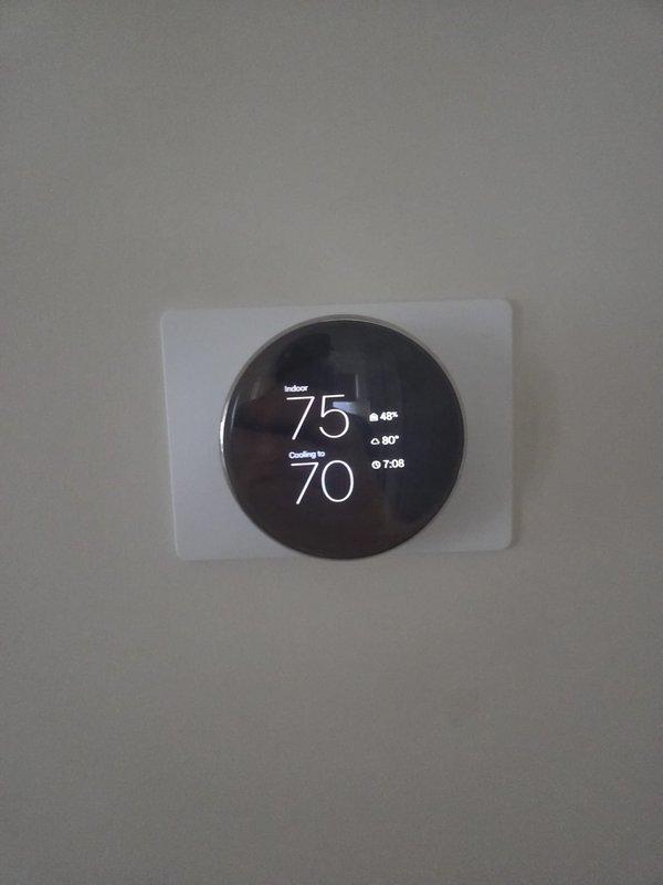 Responded to emergency no-cooling call for residential HVAC system over 10 years old. Upon arrival, found smart thermostat calling for cooling with setpoint of 70°F while indoor temperature reading showed 75°F and 45% humidity. Inspected outdoor condensing unit located on wooden platform adjacent to house with proper electrical disconnect and refrigerant line connections present. Documented system configuration and current operating conditions through diagnostic photography of both indoor thermostat display and outdoor equipment installation.