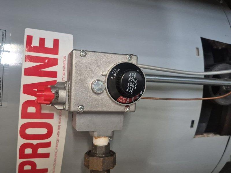 Responded to reported water heater issue on propane-fueled tanked unit. Inspected gas control valve assembly and internal combustion chamber components. Gas control valve showed proper mounting with all securing screws intact, temperature dial operational, and gas supply connection secure at brass fitting. Examined burner assembly access area and verified thermocouple wire connection. Documented equipment condition with photographic evidence showing valve assembly, propane labeling, and internal chamber access point. Unit components appeared structurally sound with normal operational wear patterns observed on exterior housing and base area.