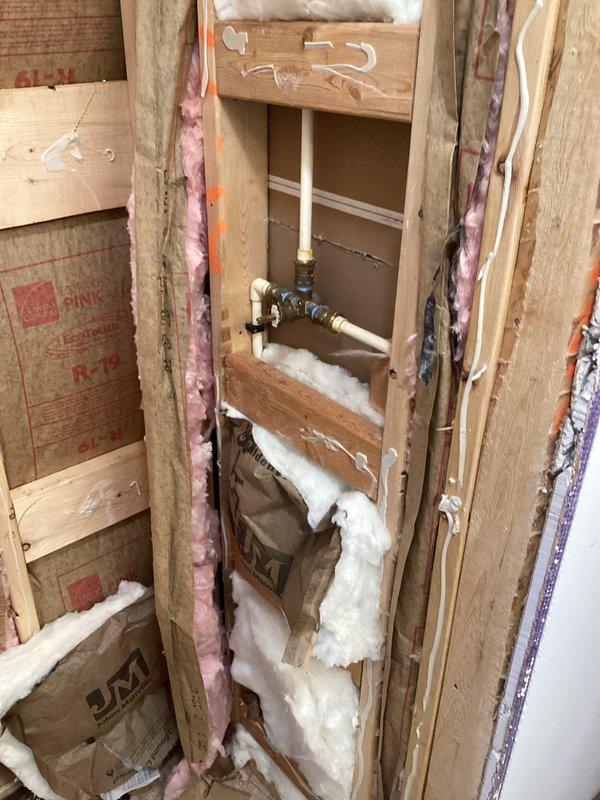Completed plumbing estimate for residential piping installation in an active construction or renovation project. Assessed exposed wall framing with vertical PVC supply lines, black iron pipes, and brass shut-off valves requiring proper insulation and finishing. Evaluated rough-in plumbing infrastructure between wall studs with existing fiberglass insulation, noting foam sealant application around pipe penetrations. Documented piping configuration, valve placement, and connection points for supply system integration. Photographed installation conditions including flooring preparation area and plumbing assembly for estimate documentation purposes.