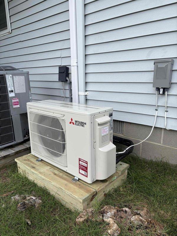 Completed installation of Mitsubishi ductless mini-split heat pump system consisting of one MITMUZ-GX12NL 12,000 BTU 25 SEER outdoor unit and one MITMSZGX12NL 12,000 BTU wall-mounted indoor air handler with remote thermostat. Routed refrigerant lines through wall and crawlspace to outdoor unit location. Mounted outdoor condenser on wooden platform adjacent to existing HVAC units. Coordinated with licensed electrician on-site to install dedicated electrical circuit and disconnect box for system power supply. System tested and operational upon completion with one-year ductless service contract included.