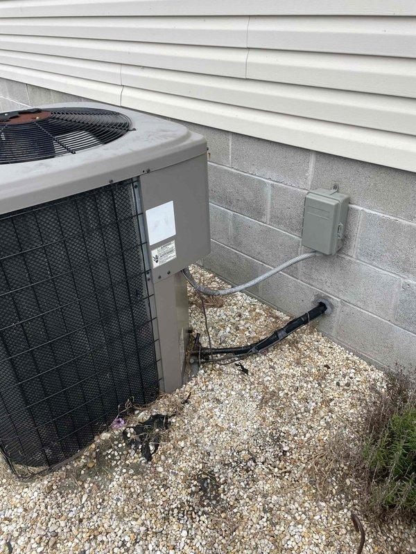 Responded to no cooling complaint with indoor temperature reading 87°F on thermostat. Upon inspection, found outdoor condensing unit operational but system failing to achieve desired cooling. Documented equipment condition with photos showing residential air conditioning condenser positioned on gravel pad against building foundation with visible refrigerant line connections and electrical disconnect. Further investigation revealed severely corroded chain component within mechanical assembly, exhibiting extensive rust and oxidation that compromised system operation. Unit scheduled for transfer to repair and replacement department for necessary component replacement and system restoration.