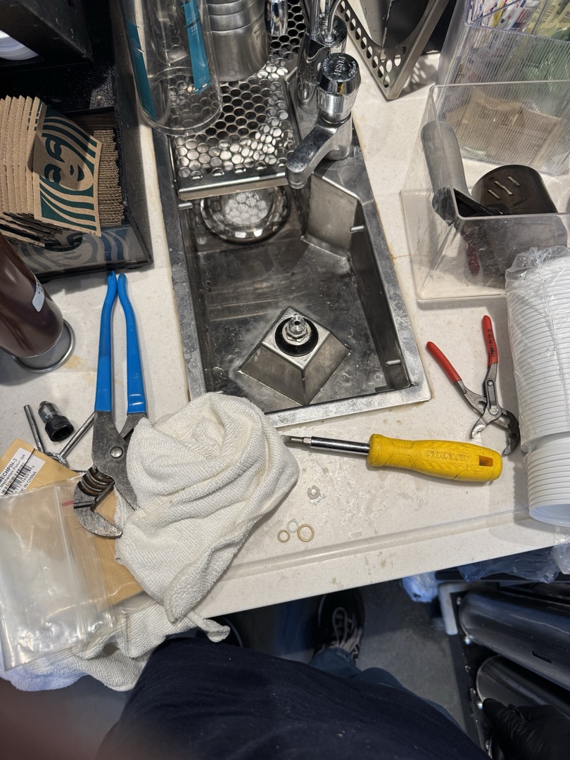 During the service, I identified an issue with the nitro brew drip tray drain line, where plastic tubing was incorrectly used instead of a 90-degree fitting, causing a kink that obstructed water drainage. Additionally, the left rhino rinser was found to be continuously spraying due to a broken bottom flat washer. I removed the rhino rinser, replaced the washer, and reset the system. I also discovered a new drip tray in a box. After removing the old tubing and tray, I installed the new drip tray and repiped using a 1” PVC female adapter, a 90-degree elbow, and 1’ of 1” PVC pipe, connected with a Fernco coupling. I tested the system by pouring water into the tray. Manager Terrence was present during the service.