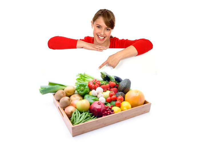 fille, jeune femme souriante pointant vers une boîte de produits frais, fruits et légumes sur un plateau en bois: haricots verts, avocats, poireau, aubergines, céleri branche, poivron rouge et vert, tomates cerises, champignons, pommes de terre, fraises, pommes, pamplemousse, citrons, grenade, baie rouge, cranberries, kiwi.