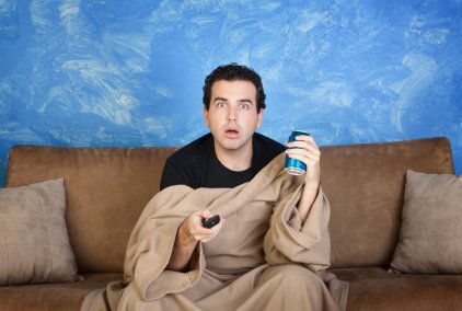 Un homme sous une couverture beige assis devant la télévision, comme hypnotisé devant un film ou une émission, la télécommande dans une main, et une boisson dans l’autre main (canette de bierre ou soda), assis sur un canapé marron.