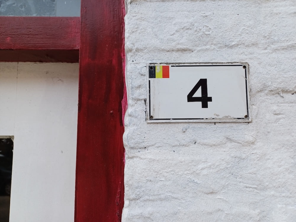 White house number plate with a small Belgian flag and the number 4 on a whitewashed wall.