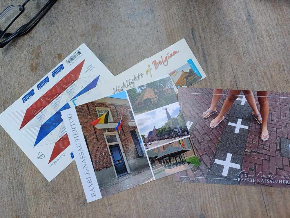 Outdoor caf&eacute; table in Baarle with drinks, pens, and a stack of postcards being written and prepared to send.