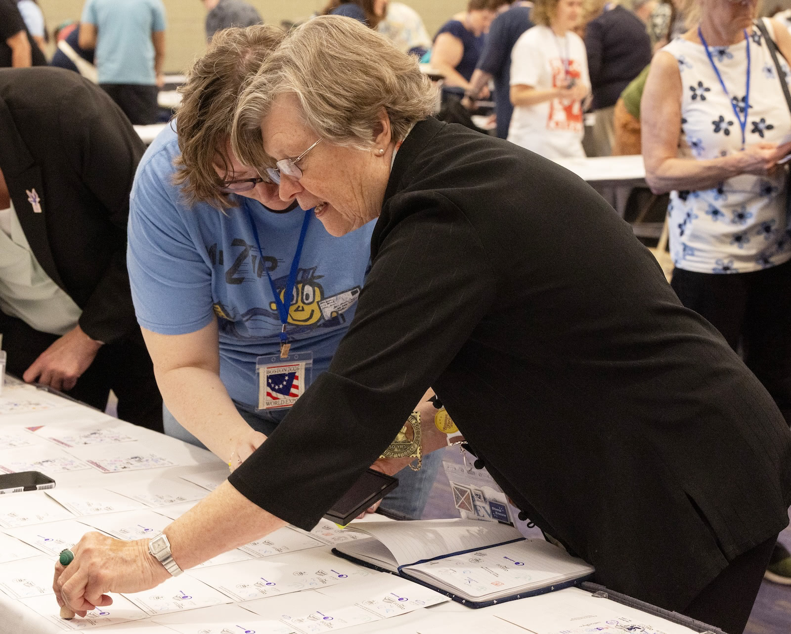 An attendee leans over a table to stamp Postcrossing stamp ceremony materials while another participant watches during the meetup after the dedication event.