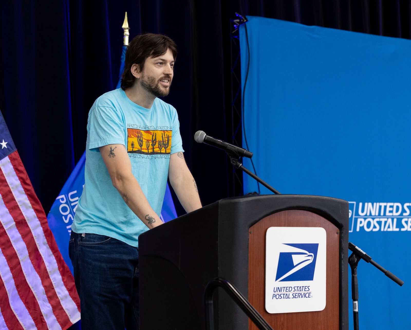 The artist Jackson Gibbs in a blue Arizona-themed shirt talks at a United States Postal Service podium during the Postcrossing stamp dedication ceremony.