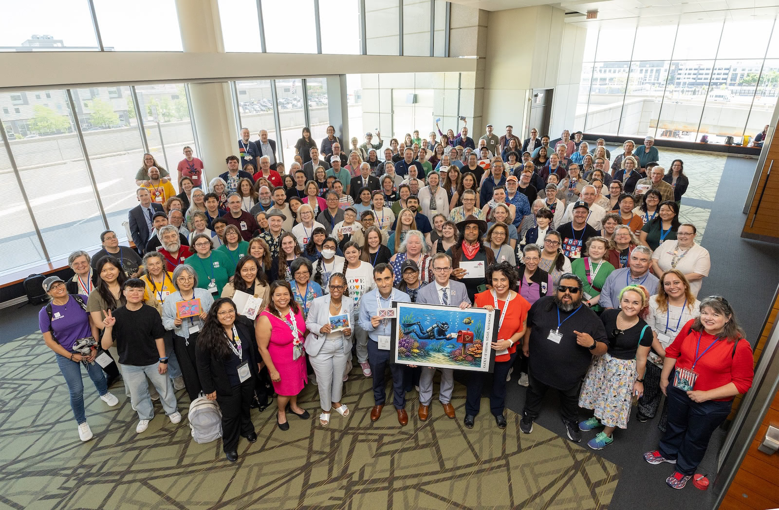 A large group of Postcrossing community members poses together after the stamp dedication ceremony, with several people holding postcards and stamp artwork.