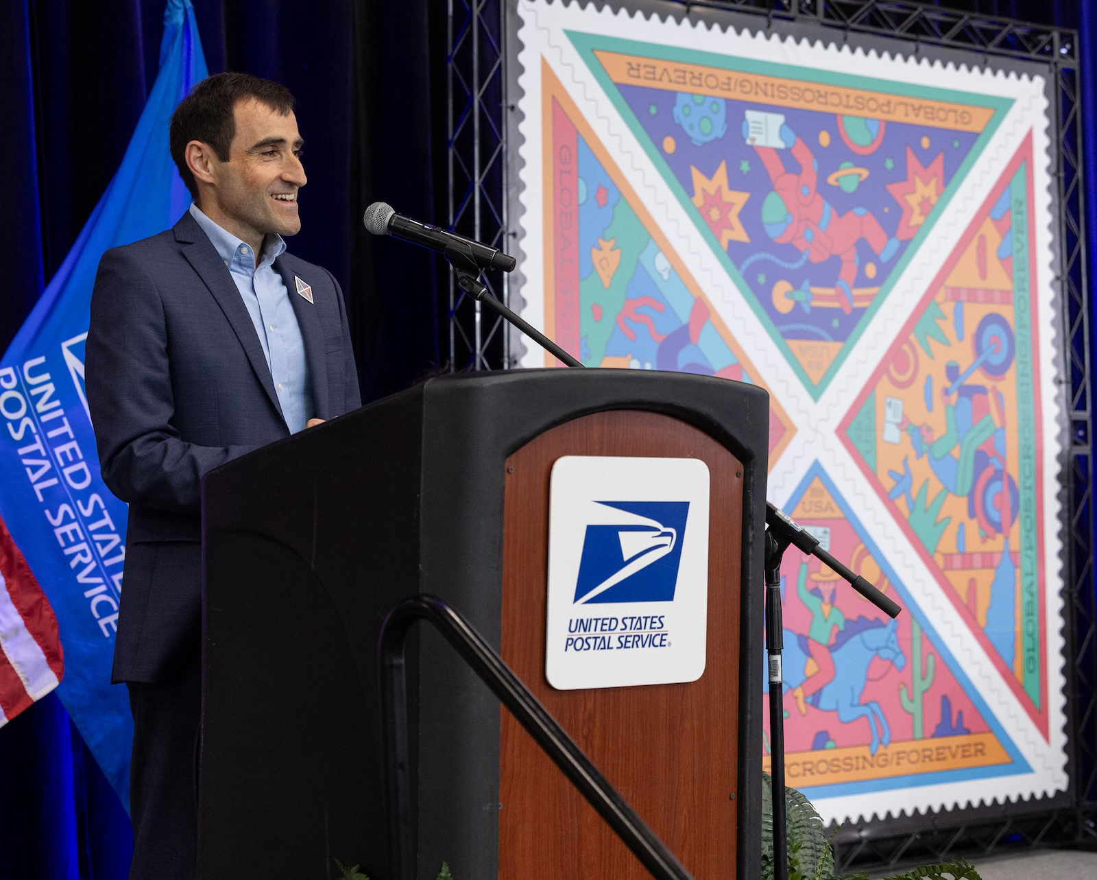 Paulo stands at a United States Postal Service podium during the Postcrossing stamp dedication ceremony, with enlarged artwork of the new triangular stamps displayed behind him.