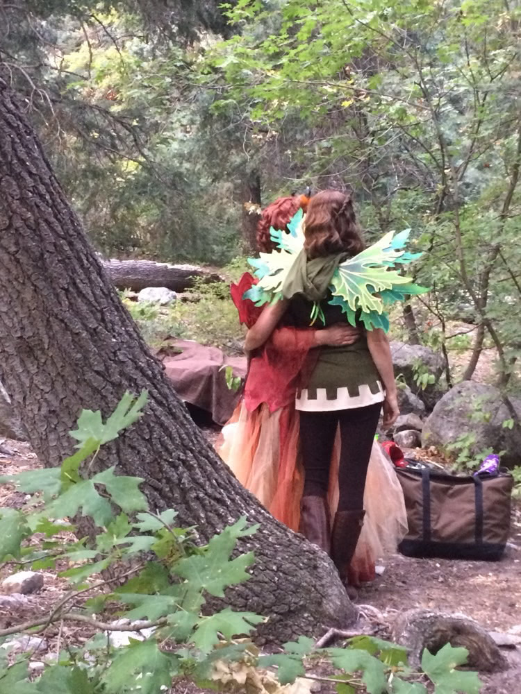 Two people dressed as fairies walk together through a forest path.