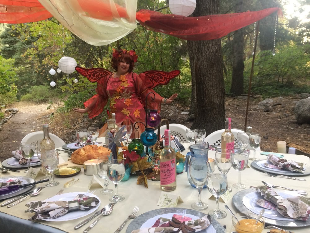 A person in a woodland fairy costume standing beside a decorated outdoor table set for a fairy tea party.