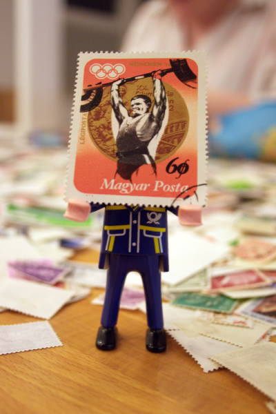 A small mail carrier toy holds a Hungarian stamp featuring an illustration of an Olympic weightlifter