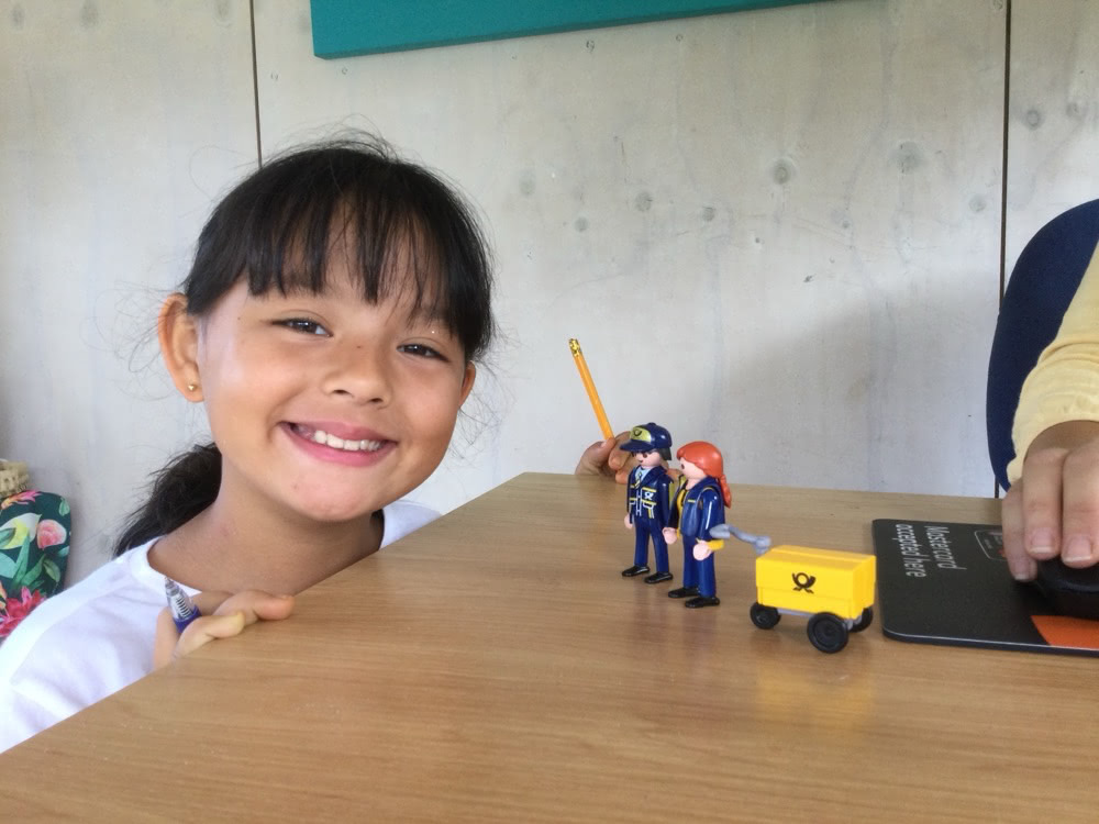 Smiling child sitting at a table beside the two Little Mail Carrier figures and their tiny yellow mail cart.